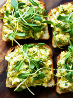 Four slices of toasted sourdough bread topped with avocado-egg salad and sprouts.