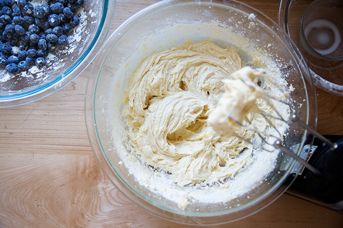An overhead shot of a bowl of blueberry muffin batter and a handheld mixer.