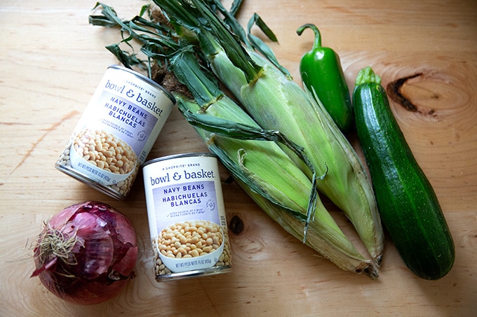 An onion, two cans of white beans, two ears of corn, one jalapeno, and one zucchini on a countertop.