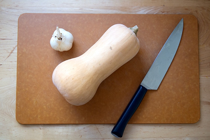 A head of garlic, a whole butternut squash, and a knife on a cutting board.