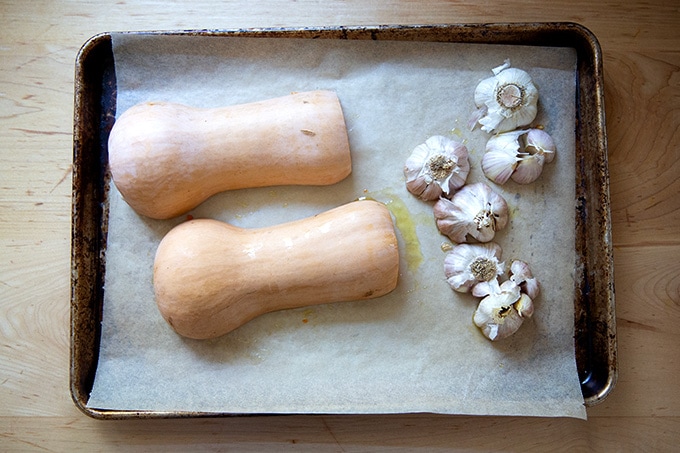 A halved butternut squash and a few halved heads of garlic on a sheet pan.