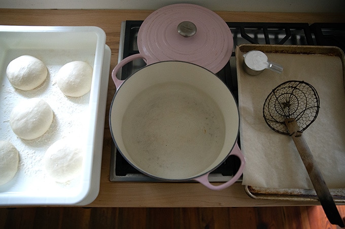 A pot of water on a stovetop aside risen pretzel roll dough balls and a sheet pan.